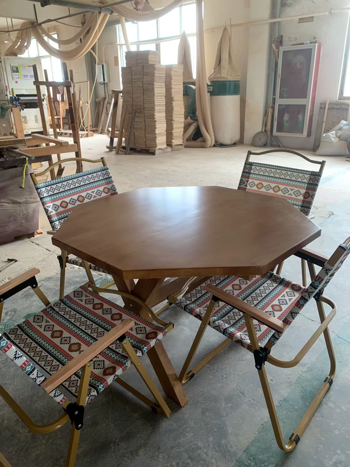 Folding camp-style table and chairs with a wood-grain top and patterned sling seats on a factory floor
