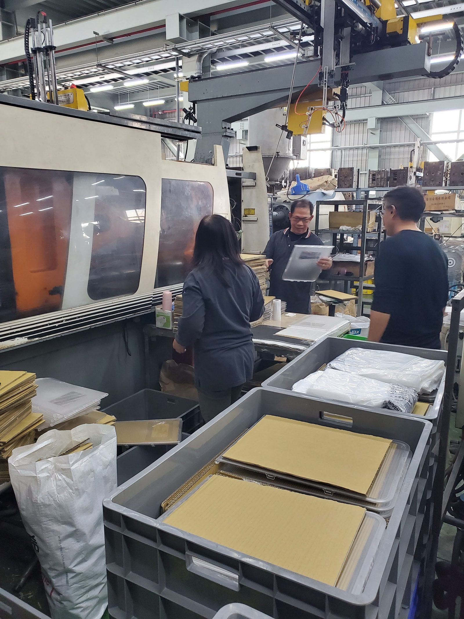 An injection molding press with a take-out robot on the gantry above it, as staff at the press hold up and check freshly molded clear parts beside crates of finished parts