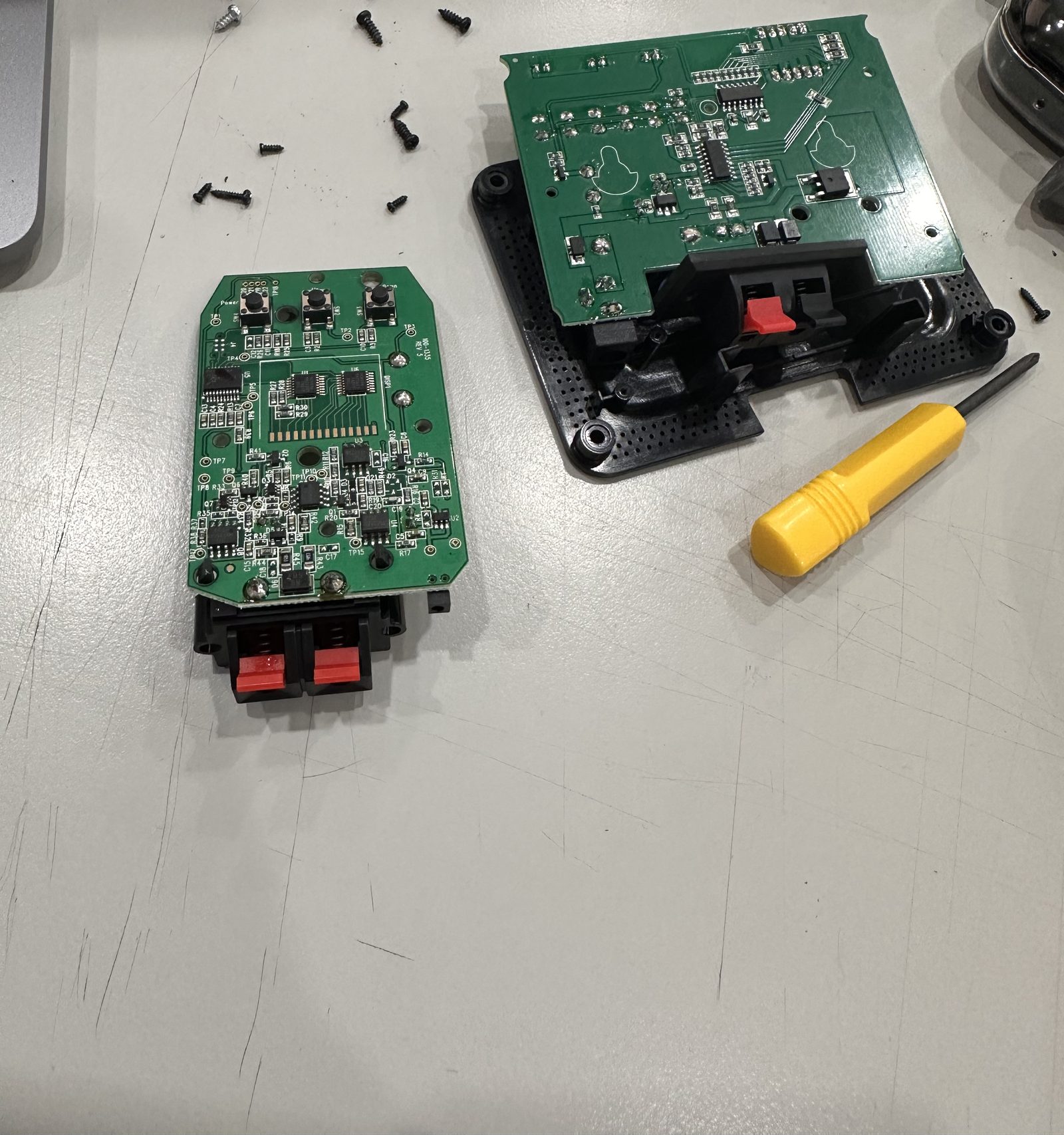Two green circuit boards taken out of their black plastic housings on a bench, with screws and a yellow screwdriver beside them