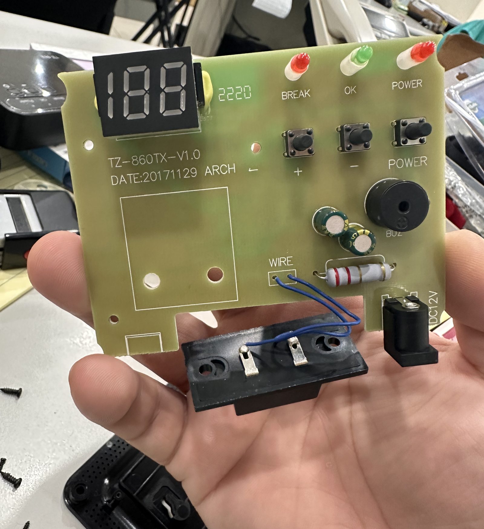 A hand holding a control circuit board with a three-digit display, buttons and a board revision and date code printed on it