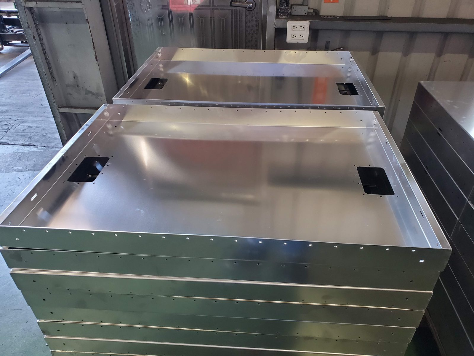 Bare sheet metal enclosure shells with punched cutouts and rows of holes stacked on a factory floor