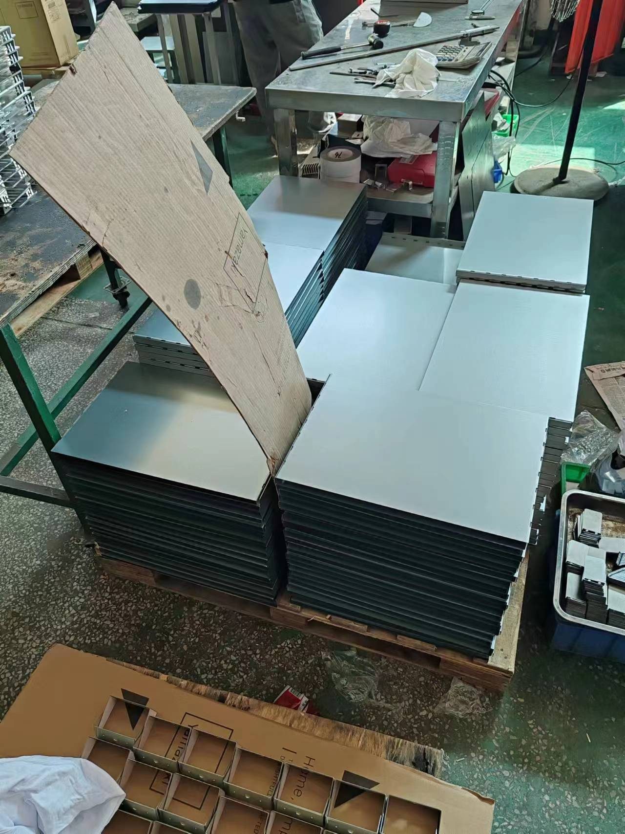 Stacks of flat sheet-metal panels on a wooden pallet on a factory floor, with a carton cover folded back