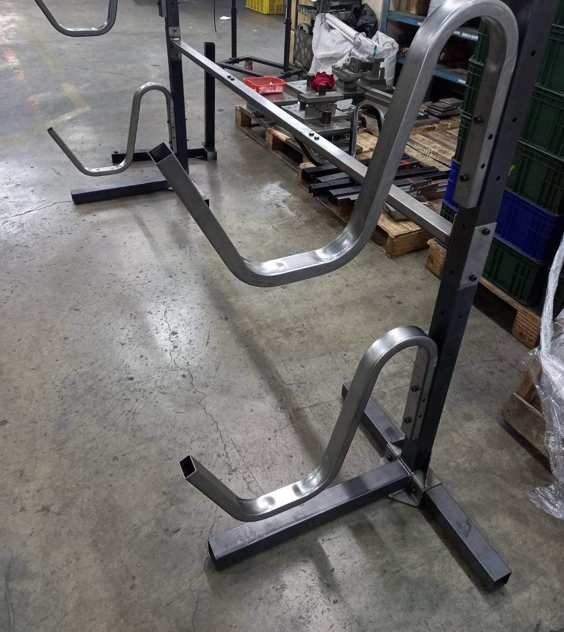 A first-build stand of bent stainless steel tube on a black steel frame, standing on a factory floor