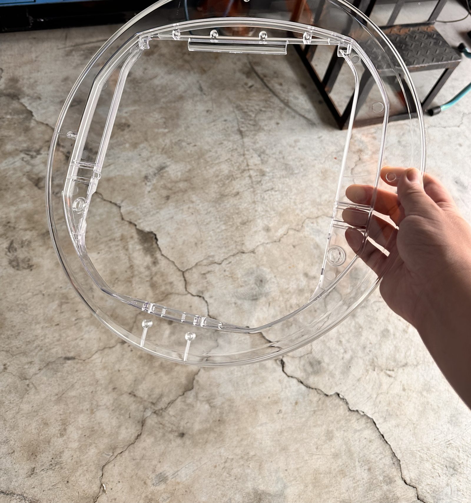 A hand holding a clear plastic molded frame with screw bosses and clips over a concrete factory floor