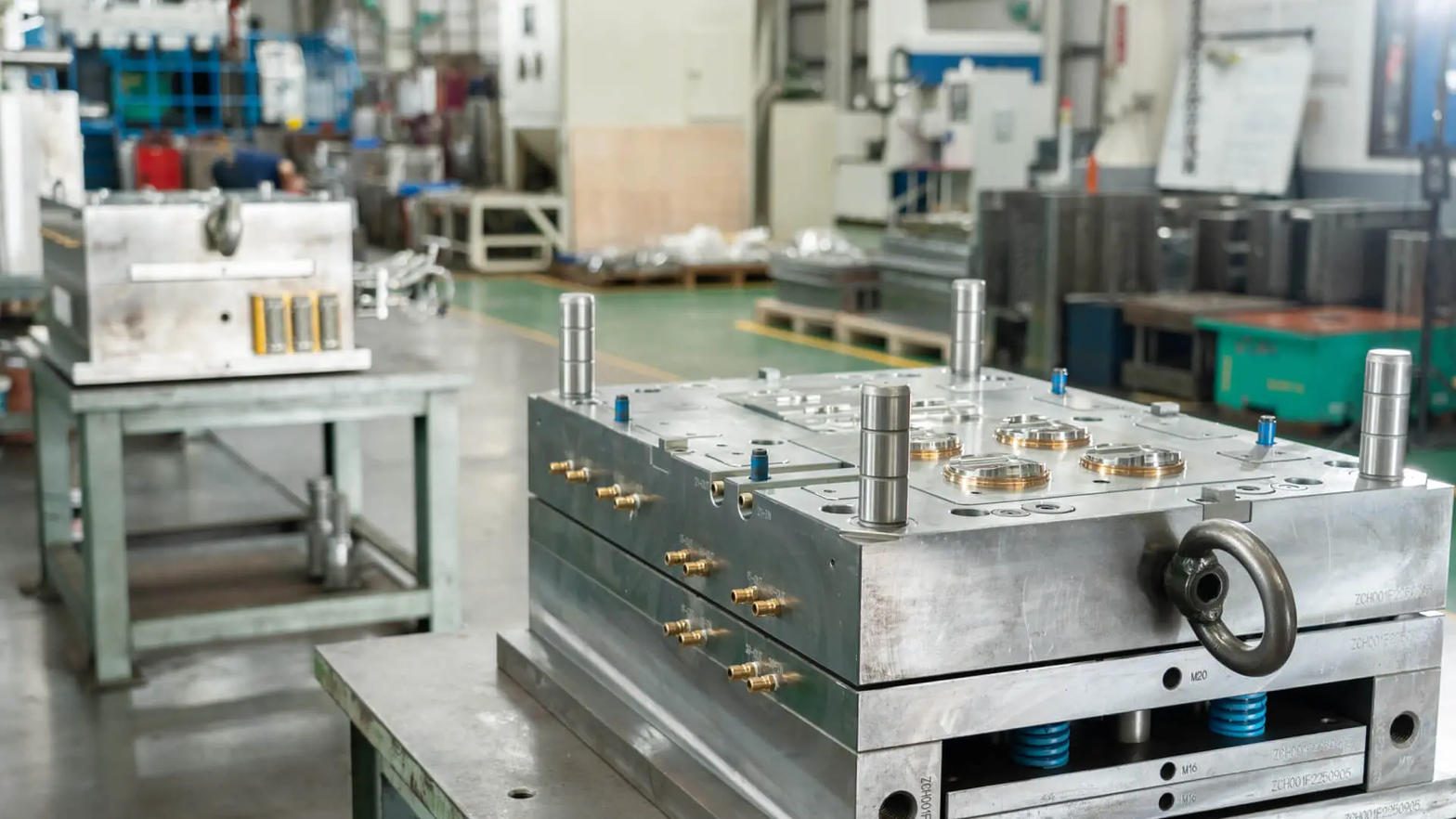 A closed injection mold base on a tool-room bench, with tall guide pins on top, brass cooling-water fittings along the side and blue springs in the ejector housing, and a second mold on a stand behind it