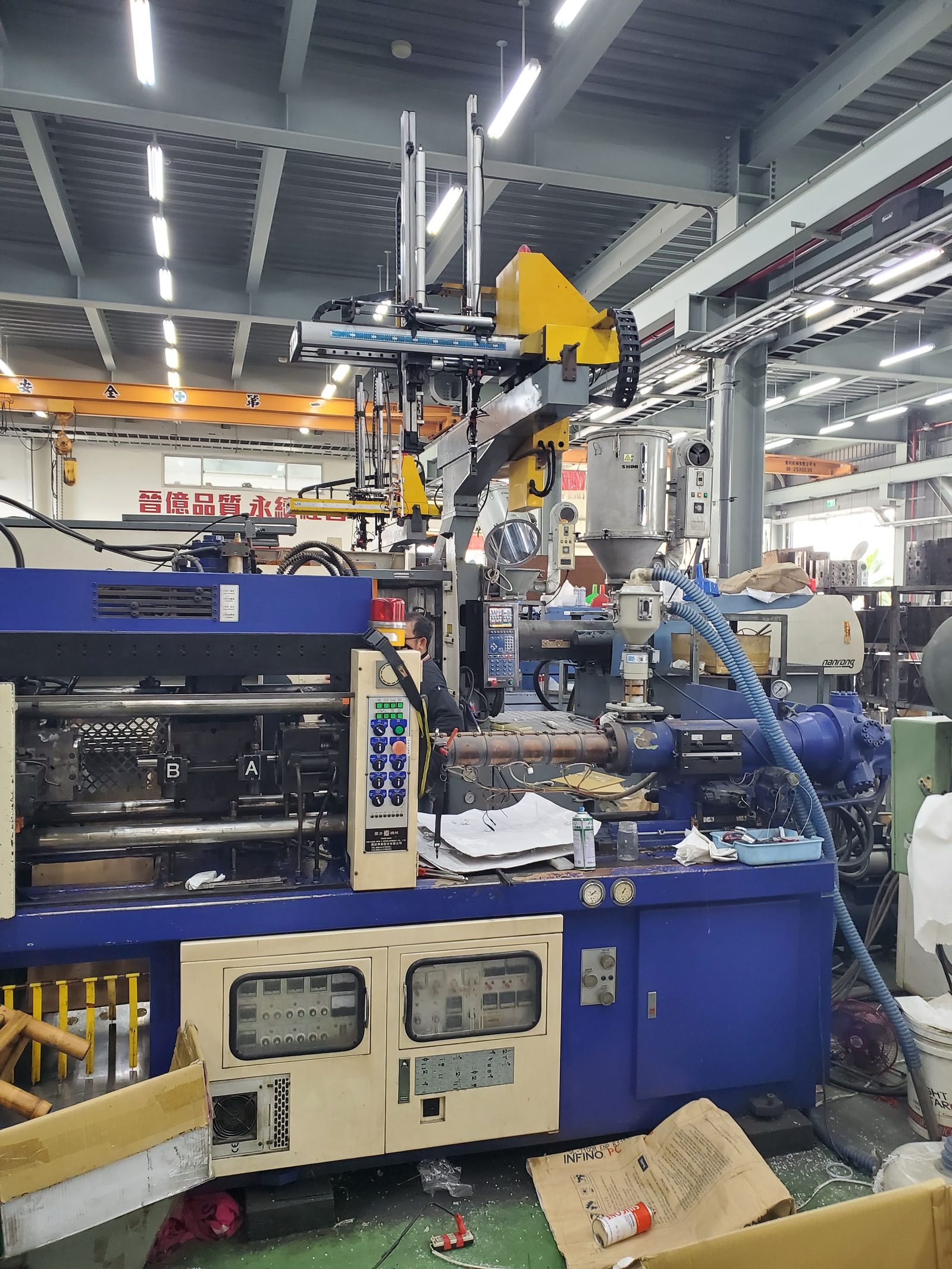 An injection molding press on a factory floor with a take-out robot mounted above the platen and a resin dryer beside it
