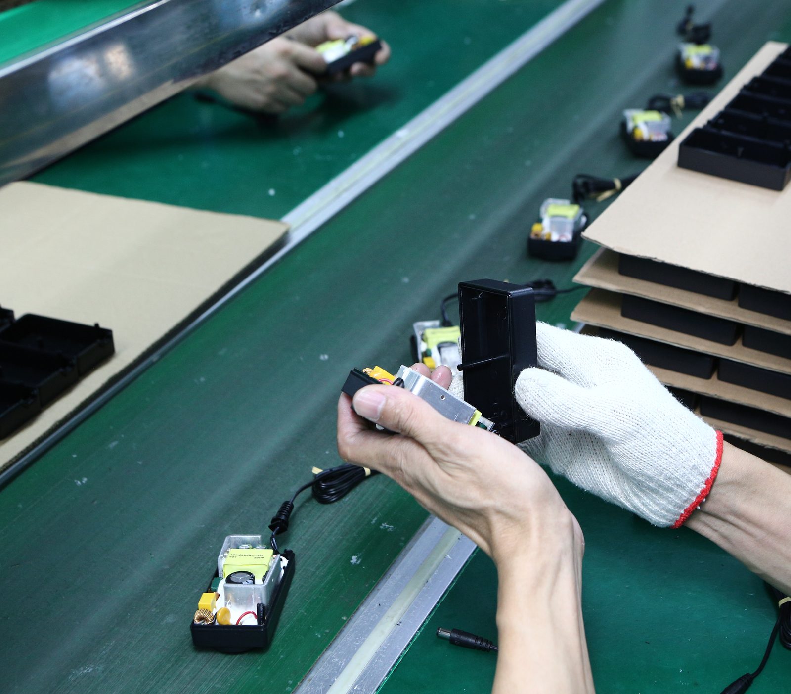 A gloved hand holding a black plastic housing while the other hand fits a small circuit board into it on a green assembly line, with stacked trays behind