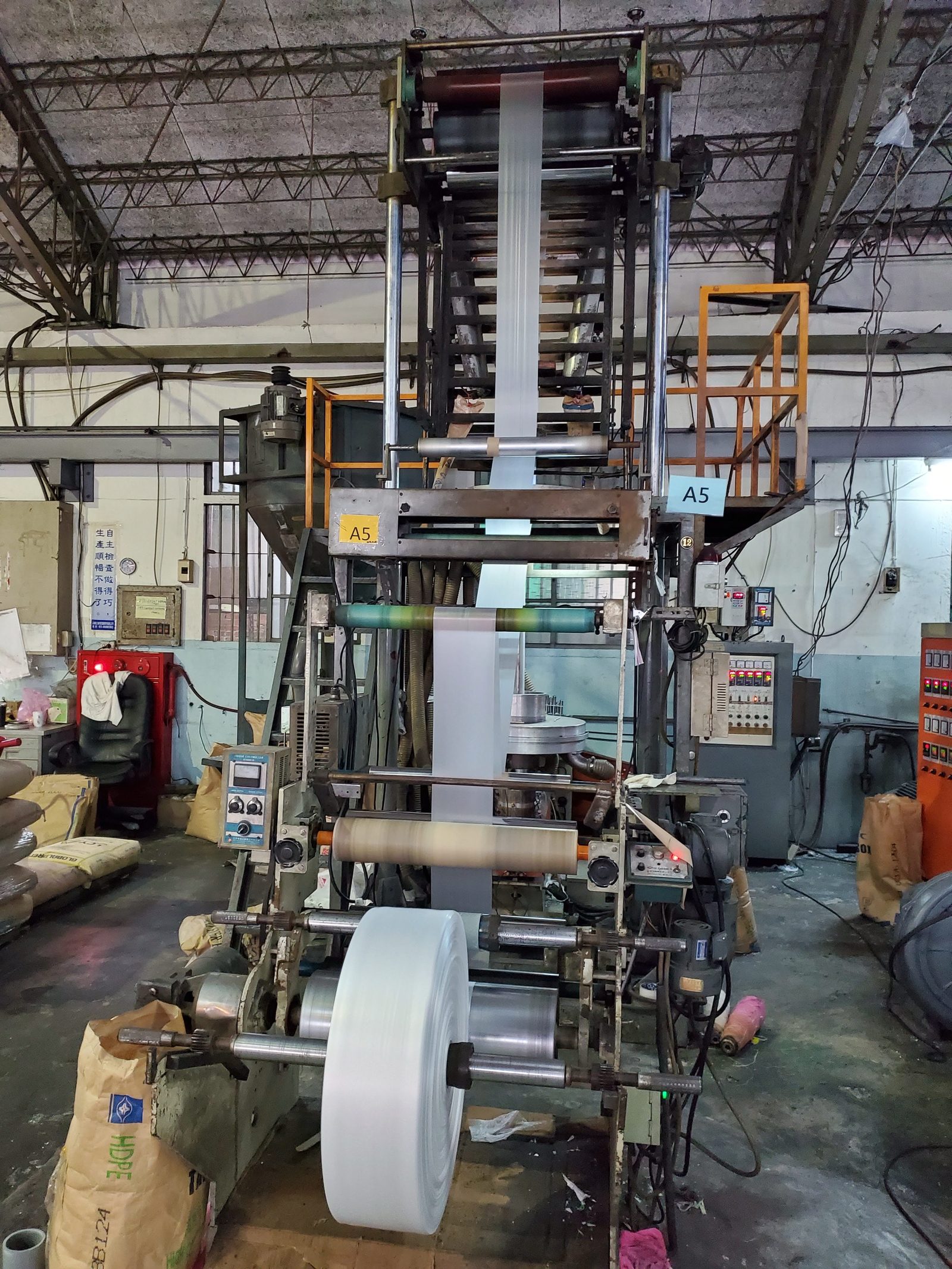 A blown film extrusion tower pulling a tube of white HDPE film up to the nip rolls, with a finished roll at its base and resin sacks beside it