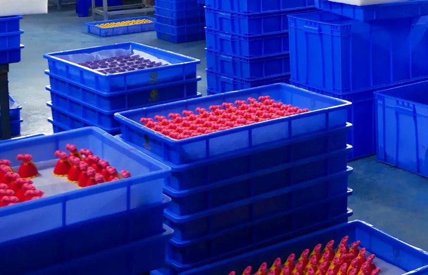 Blue plastic trays stacked on a factory floor, filled with rows of small red and purple molded plastic parts