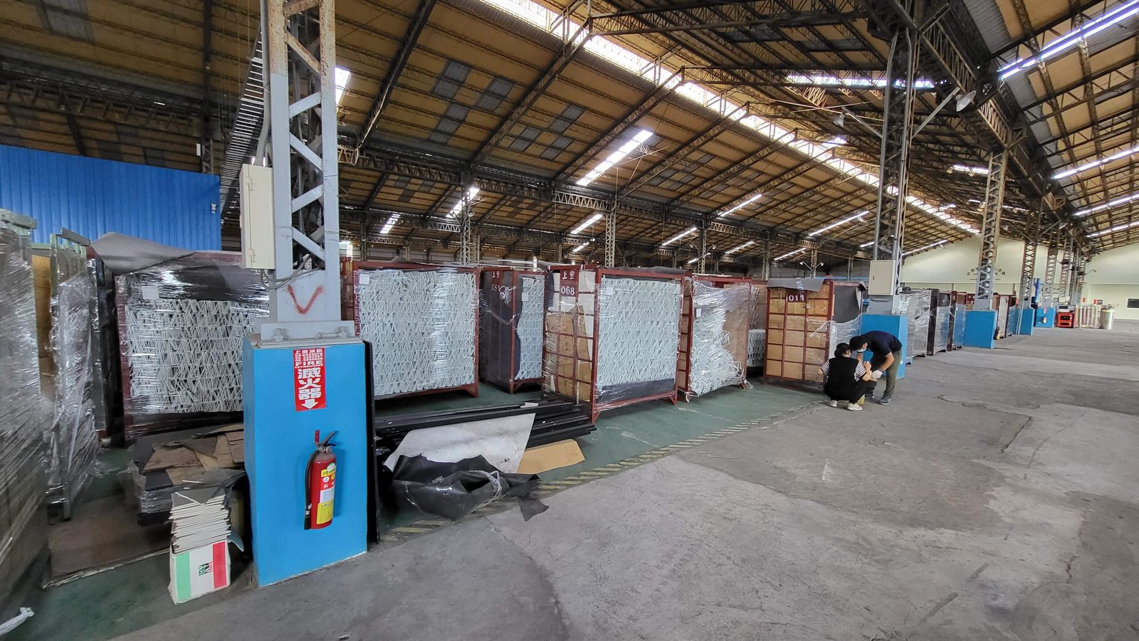 A long factory warehouse aisle with steel frames shrink-wrapped in red racks, staged before loading