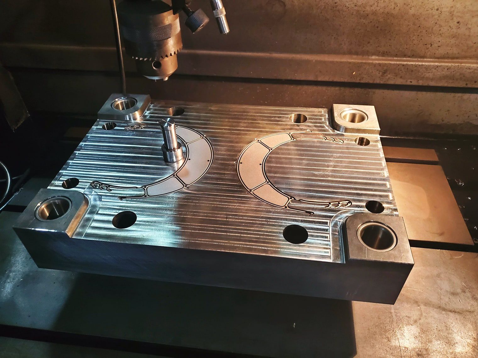 A machined steel mold plate with curved cavity detail and guide bushings, on a machine table