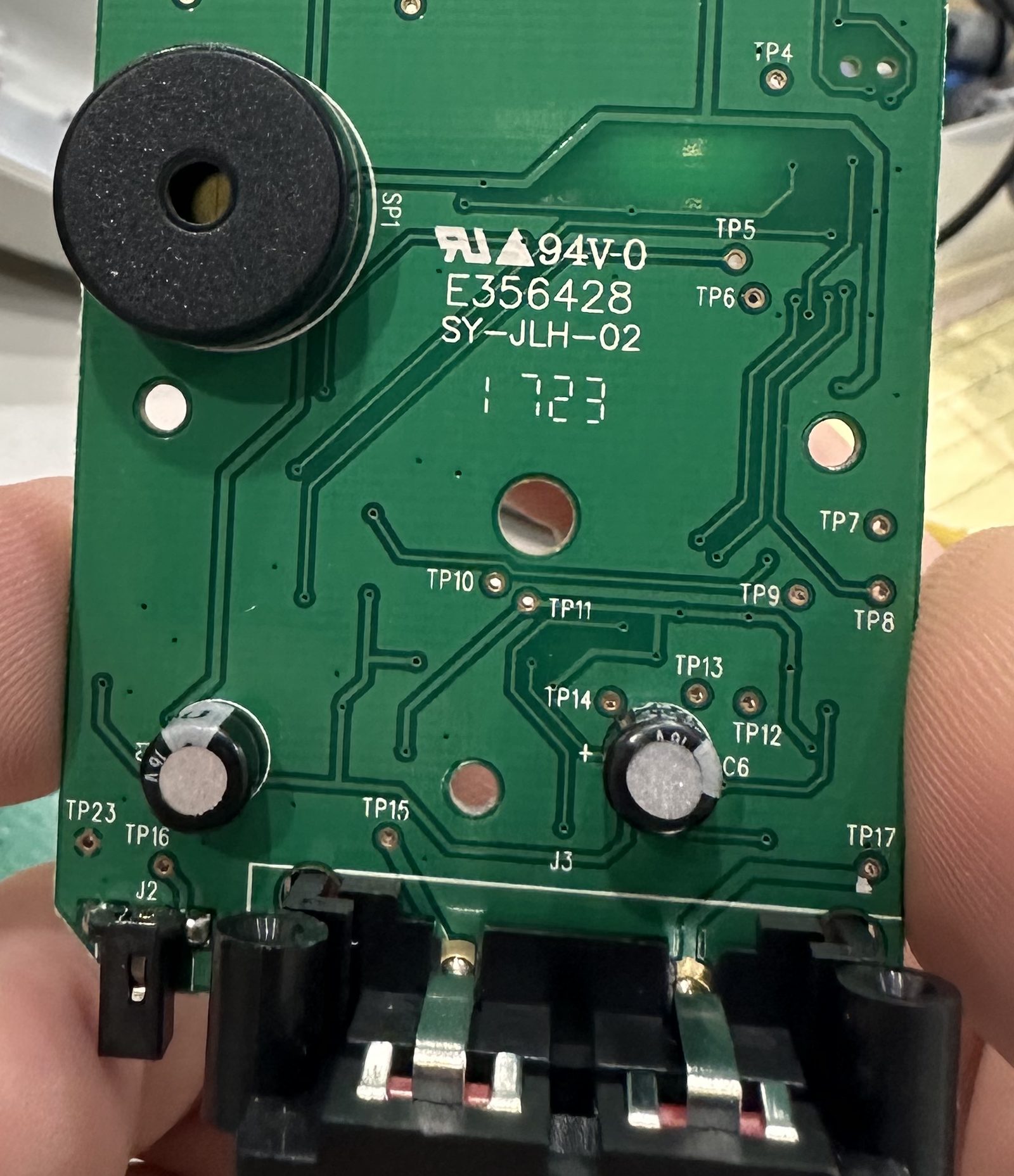 The back of a green circuit board held between two fingers: a round black buzzer, two can-shaped capacitors, a black connector along the bottom edge and labeled test points