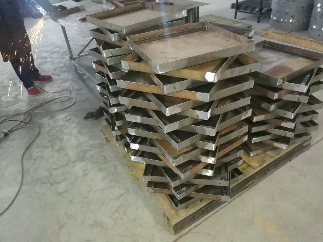 Stacks of welded stainless steel trays on a pallet while a worker grinds seams, the work that decides country of origin