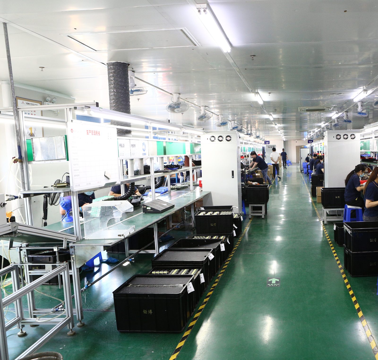 A long electronics assembly line with operators at green-topped stations and crates of boards