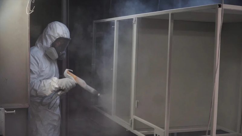 An operator in a full suit sprays powder onto a large metal cabinet inside the booth.