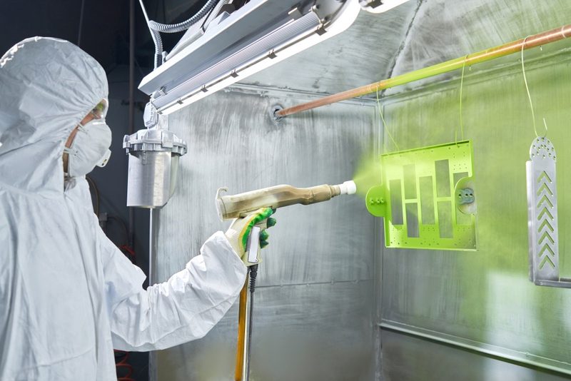 A punched panel receives green powder, with even coverage on the cutouts and edges. An uncoated part hangs beside it.