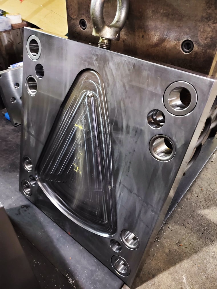 A steel mold plate with a triangular cavity, lifted on an eyebolt